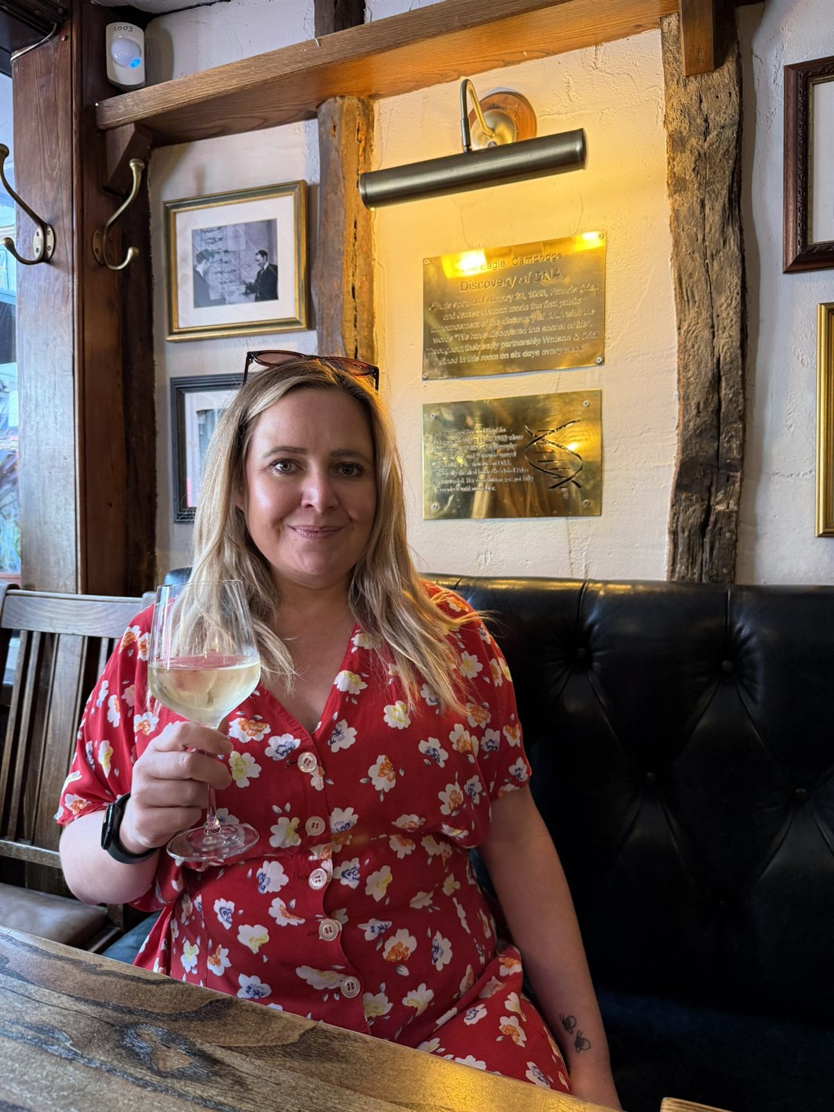 Kat on the bench at the Eagle pub in Cambridge with a glass of white wine, the DNA plaque lit on the wall behind her
