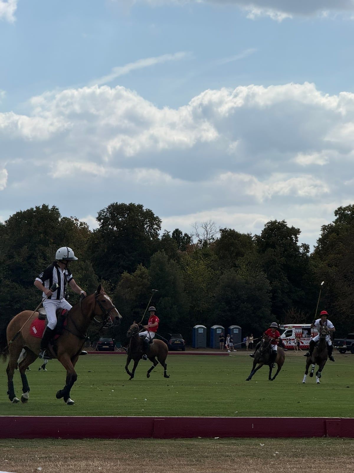 A mounted umpire in stripes and players in red mid-chukka at Ham Polo Club