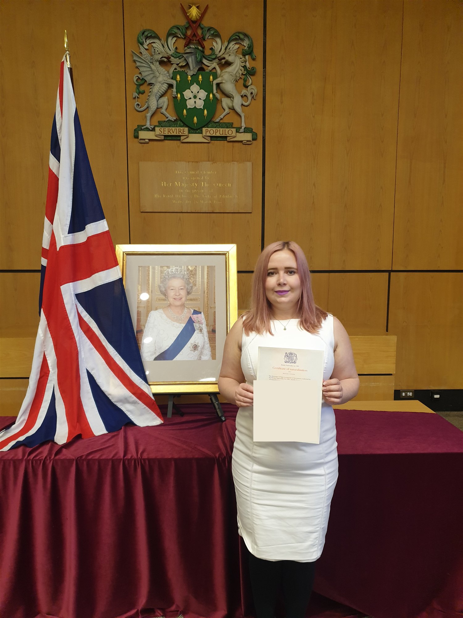 Kat in a white dress holding her certificate of naturalisation in the council chamber at Bromley Civic Centre, 22 October 2019, next to the Union flag and a portrait of the Queen.