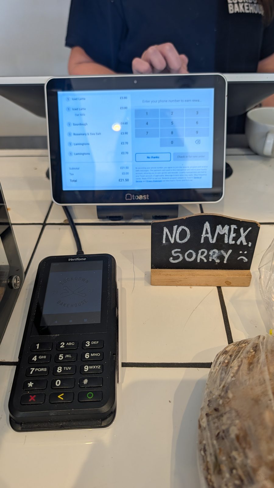 A bakery counter till showing a £21.50 order beside a chalkboard reading no Amex sorry
