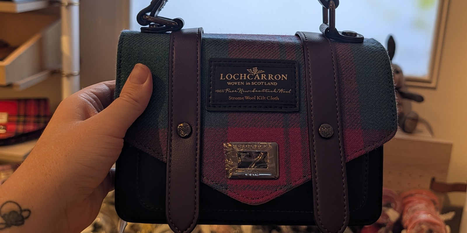 Kat's finished Islander mini bag with a pink tartan lid and purple straps, held up in front of The Workshop sign